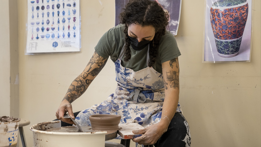 A masked student uses a pottery wheel